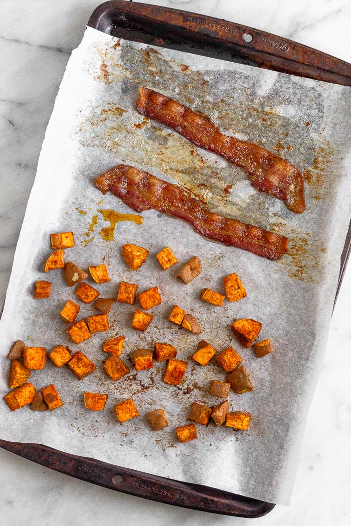 Overhead shot of a baking sheet lined with parchment paper with diced sweet potato and two pieces of bacon.