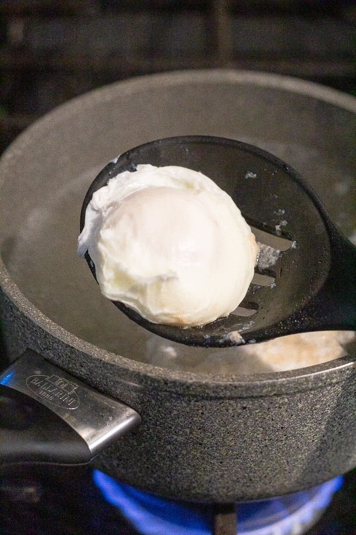 Close up of slotted spoon holding a poached egg overtop a sauce pan.