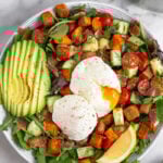 Overhead shot of a breakfast salad with veggies, sliced avocado, and two poached eggs with one of them cut open and the yolk is gushing out.