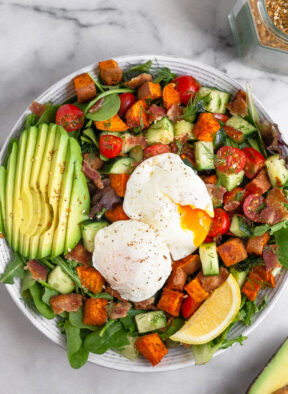 Overhead shot of a breakfast salad with veggies, sliced avocado, and two poached eggs with one of them cut open and the yolk is gushing out.