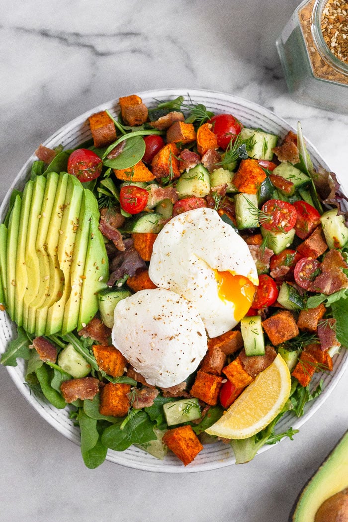 Overhead shot of a breakfast salad with veggies, sliced avocado, and two poached eggs with one of them cut open and the yolk is gushing out.