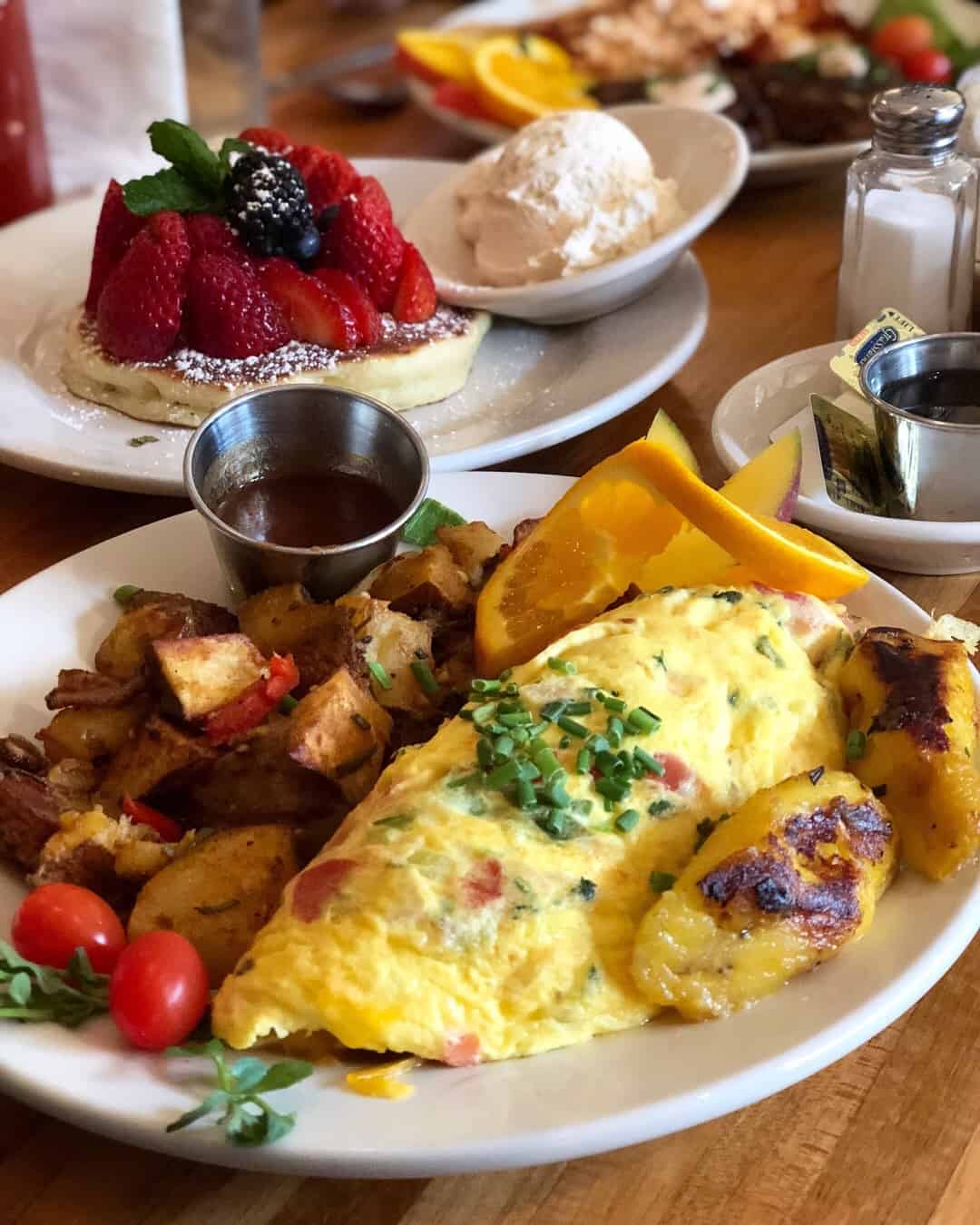 Loaded veggie omelet wit h a side of crispy potatoes and plantains with a pancake topped with berries and a side of whipped cream behind it
