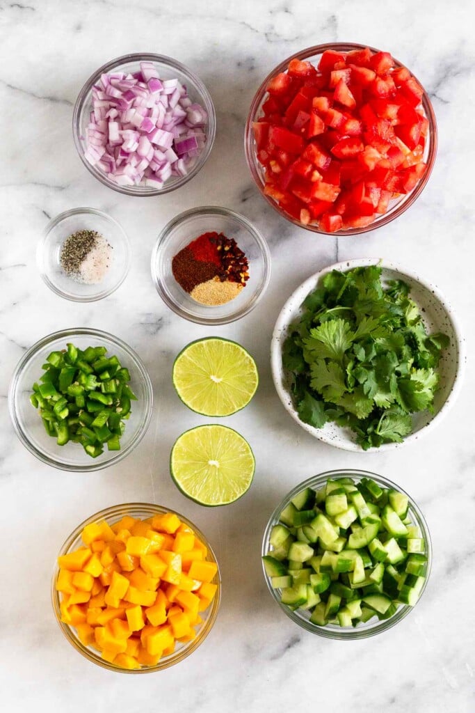 White marble counter with a bowl of diced tomatoes, a bowl of spices, a plate of fresh cilantro, a bowl of diced cucumbers, a bowl of diced mango, a lime cut in half, a bowl of diced jalapeños, a bowl of salt and pepper, and a bowl of diced red onion.
