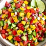 Mango cucumber salsa in a white bowl garnished with two lime wedges. The bowl is sitting on a large plate with tortilla chips on it.
