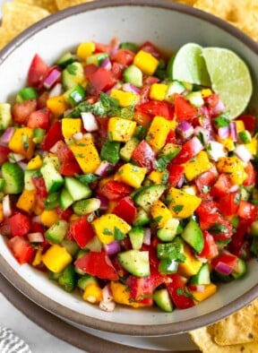 Mango cucumber salsa in a white bowl garnished with two lime wedges. The bowl is sitting on a large plate with tortilla chips on it.