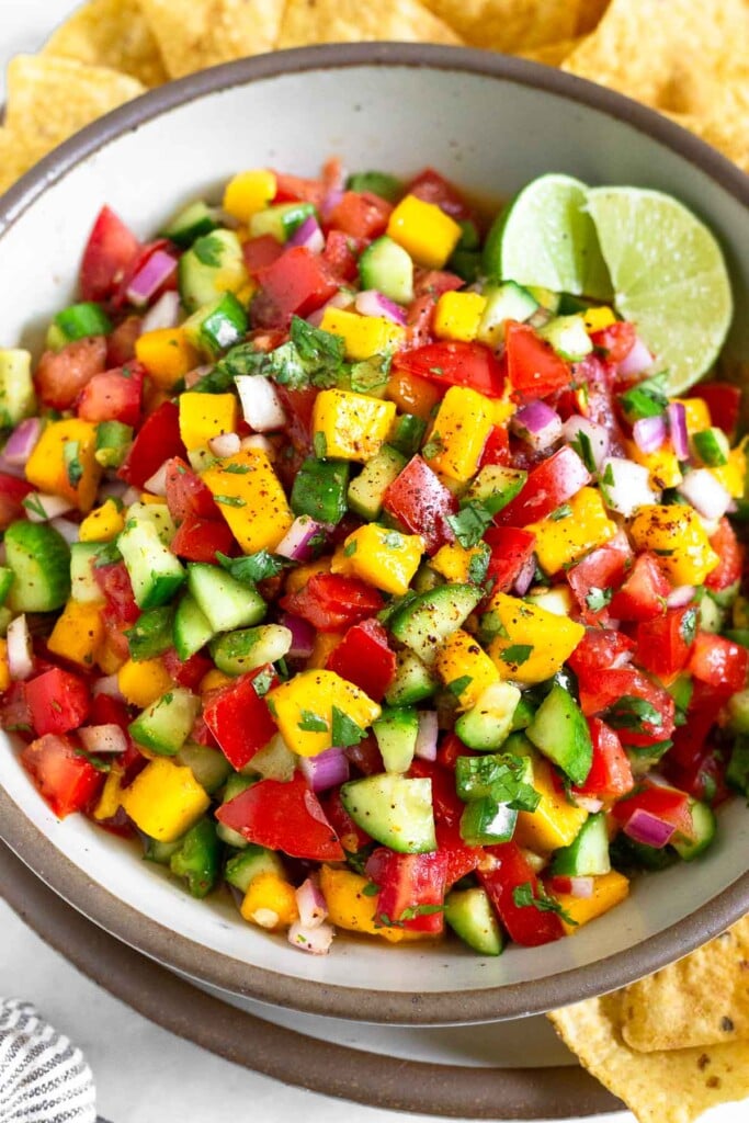 Mango cucumber salsa in a white bowl garnished with two lime wedges. The bowl is sitting on a large plate with tortilla chips on it.