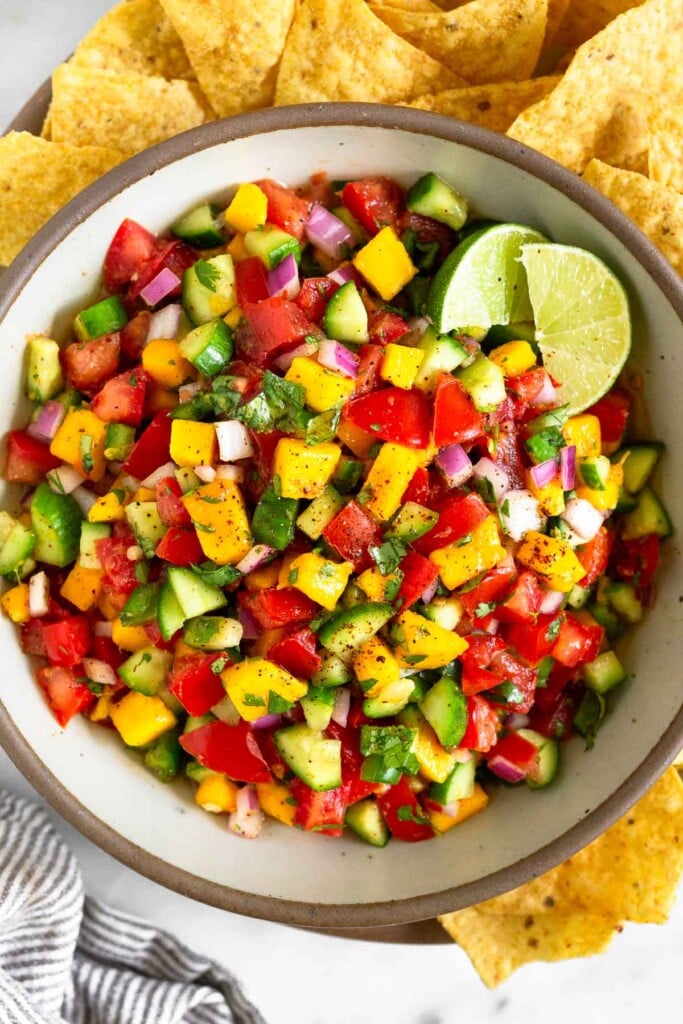 Fresh mango salsa with tomatoes, cucumber, red onion, and cilantro in a large white bowl garnished with 2 lime wedges. Around the bowl are tortilla chips.