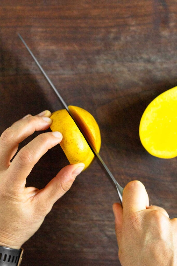 A mango being held up on a cutting board with a knife cutting off one of the sides. The other side is already cut off.