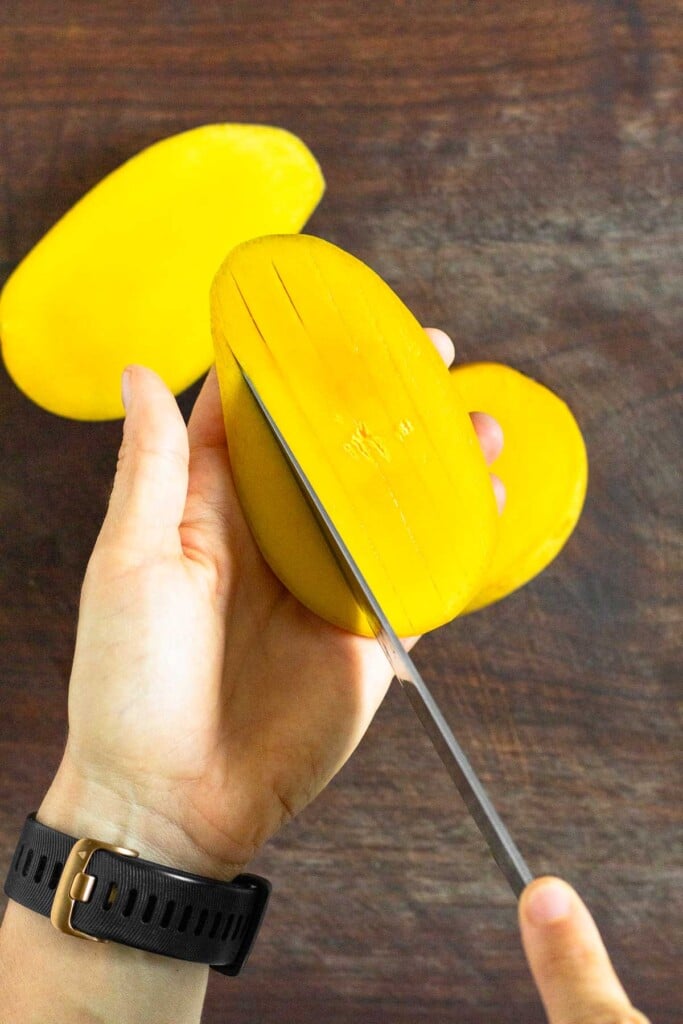 Half a mango being held over a cutting board. The flesh is being sliced.