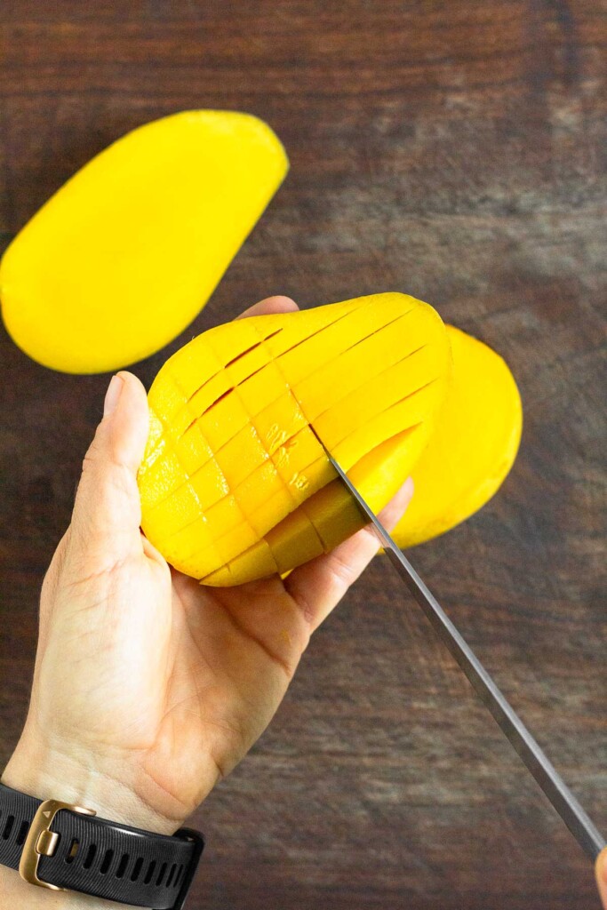 Half a mango being held over a cutting board. It is being sliced in a grid pattern.