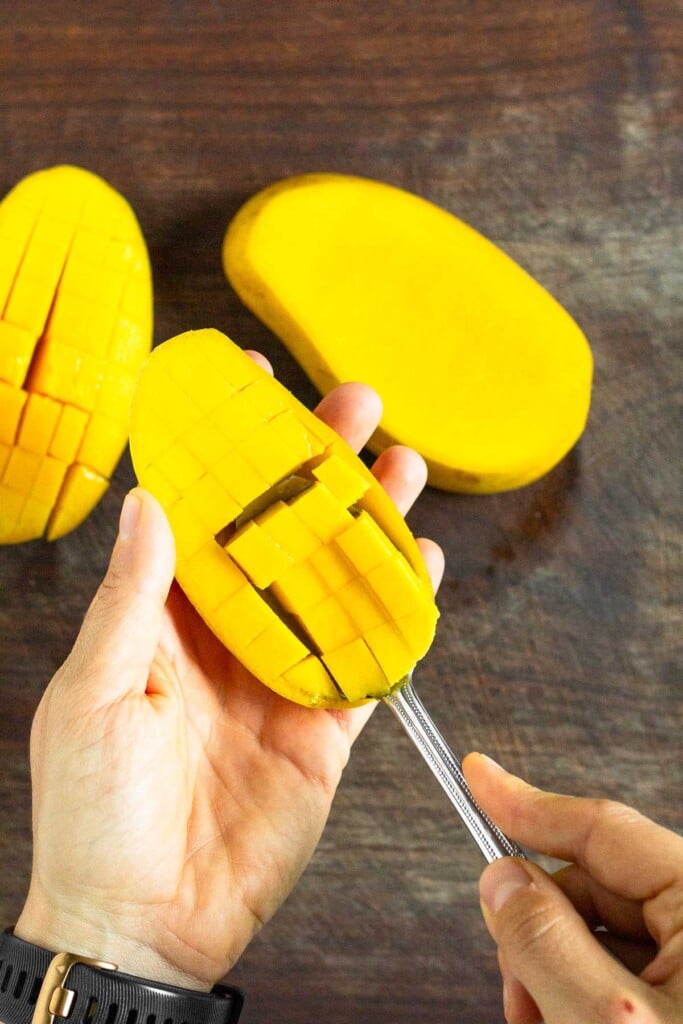 Half a mango being held over a cutting board. It has been diced and a spoon is scooping out the flesh from the skin. On the cutting board is the other half and the middle with the seed.