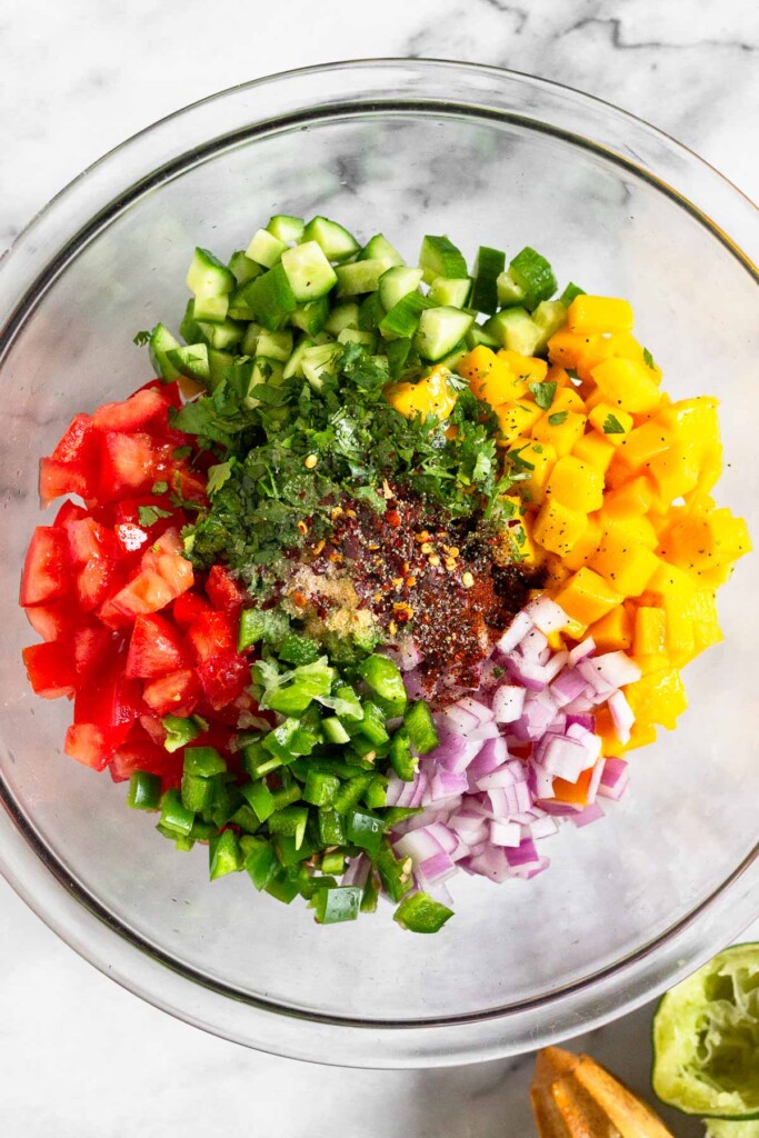 Large glass bowl with diced mango, diced red onion, diced jalapeño, diced tomatoes, diced cucumber, cilantro, and spices before it is mixed together. Next to the bowl is half a juiced lime and a wooden reamer.