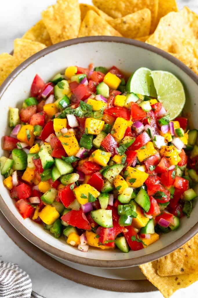 Mango salsa with tomatoes, cucumbers, red onion, and cilantro in a white bowl. It is garnished with 2 limes wedges. The bowl is sitting on a plate with tortilla chips on it.