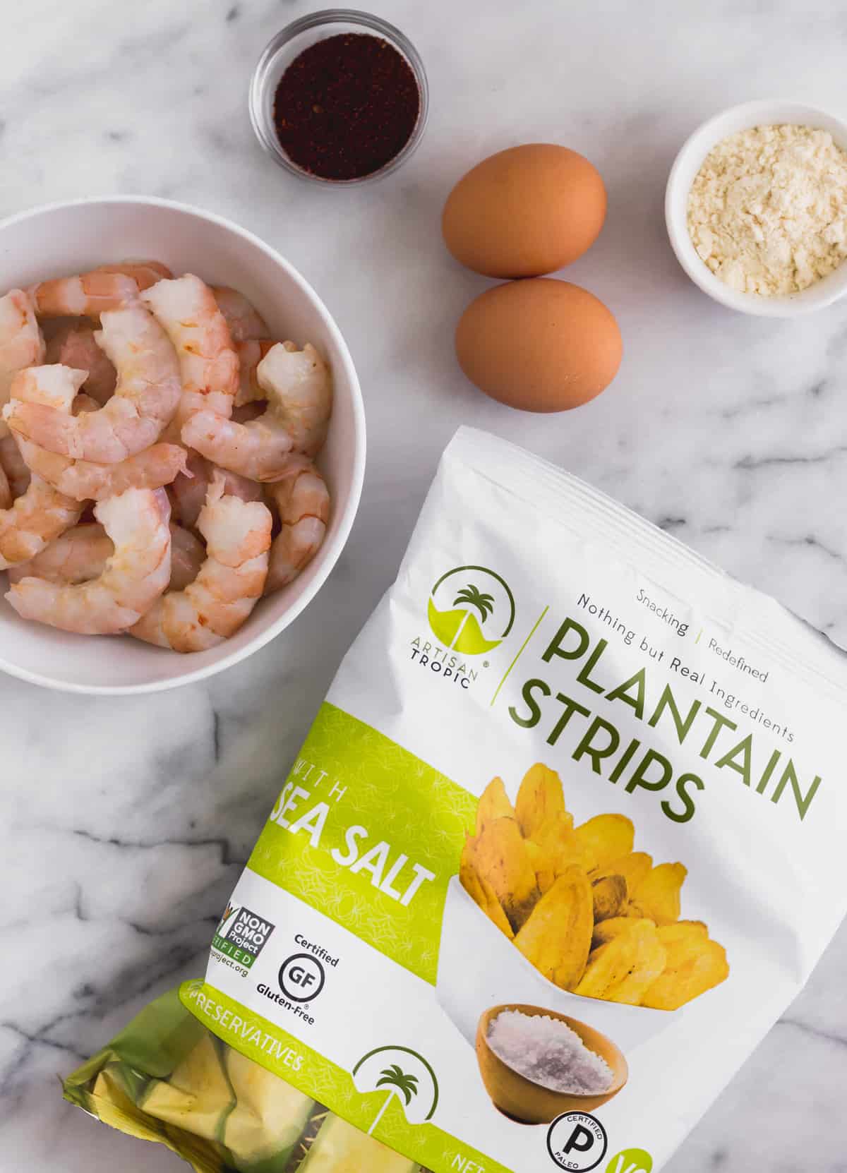 White marble counter top with a bag of plantain chips, raw shrimp, taco seasoning, eggs, and coconut flour on it