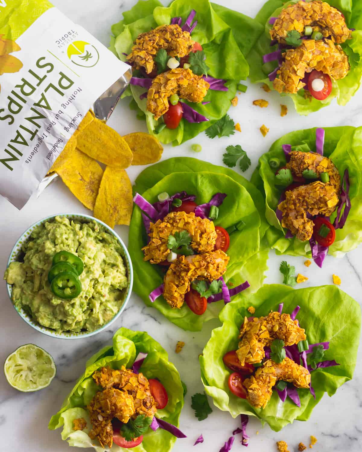 A bunch of Paleo Plantain Crusted Crispy Shrimp Tacos on a white counter with a bowl of guacamole and a bag of plantain chips around them