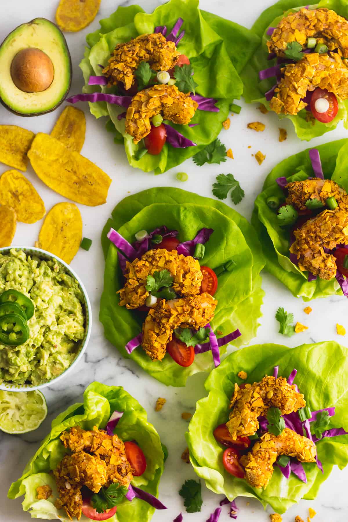 A bunch of Paleo Plantain Crusted Crispy Shrimp Tacos on a white counter with a bowl of guacamole and plantain chips around them