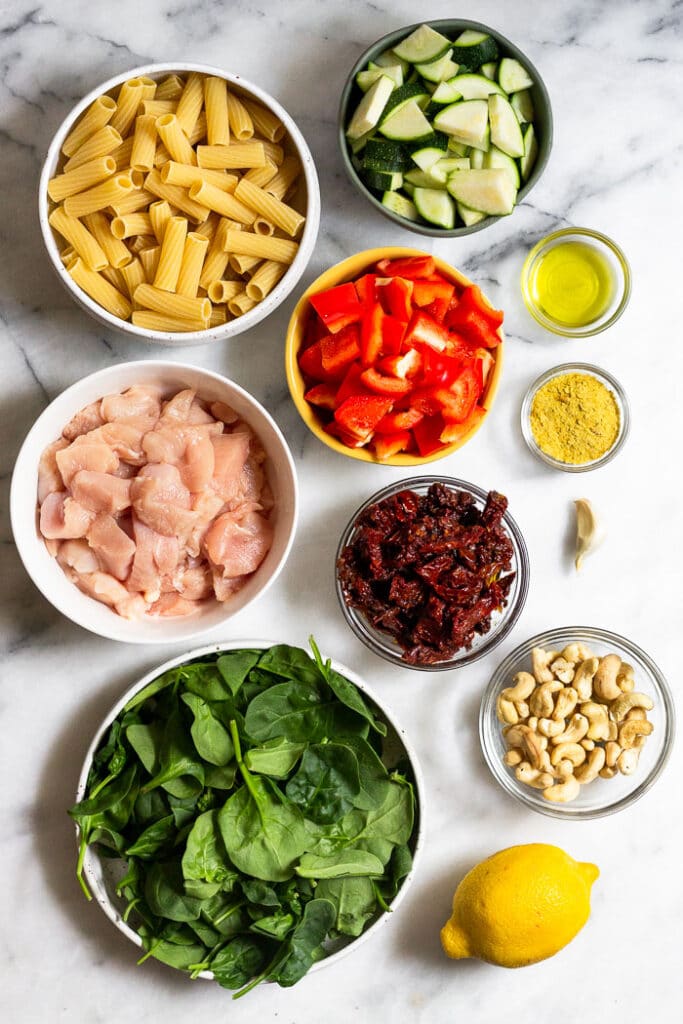 Overhead shot of a bowl of uncooked pasta, bowl of diced zucchini, bowl of olive oil, bowl of nutritional yeast, a clove of garlic, bowl of cashews, bowl of sun-dried tomatoes, bowl of diced bell pepper, bowl of raw diced chicken, and a bowl of spinach.