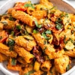 Sun dried tomato chicken pasta in a bowl topped with basil and parmesan cheese. Behind it is another bowl of pasta and bowl of sun-dried tomatoes.