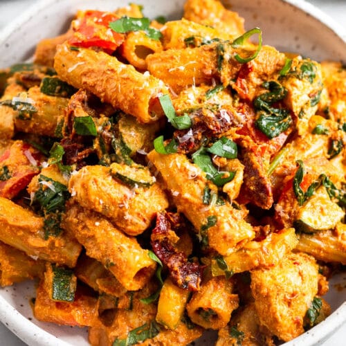 Sun dried tomato chicken pasta in a bowl topped with basil and parmesan cheese. Behind it is another bowl of pasta and bowl of sun-dried tomatoes.
