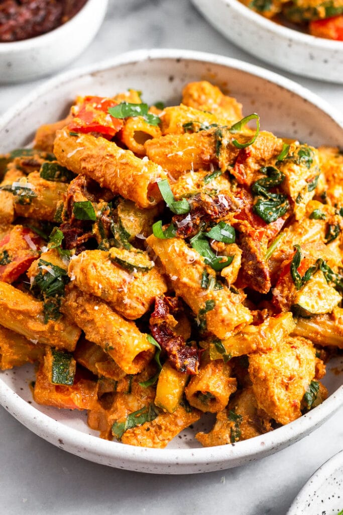 Sun dried tomato chicken pasta in a bowl topped with basil and parmesan cheese. Behind it is another bowl of pasta and bowl of sun-dried tomatoes.