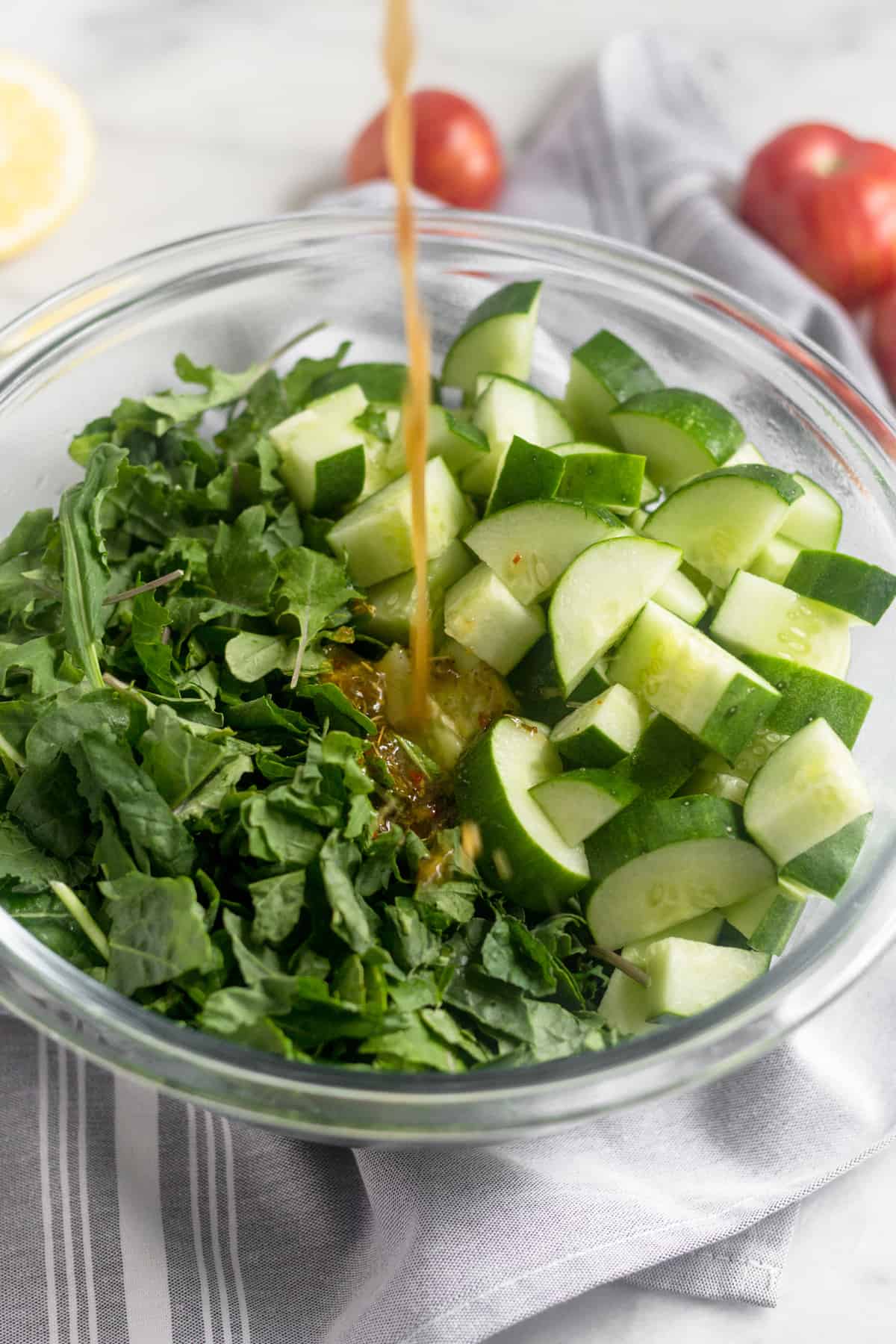 Glass bowl of chopped kale and cucumbers with a homemade Italian dressing being poured on it