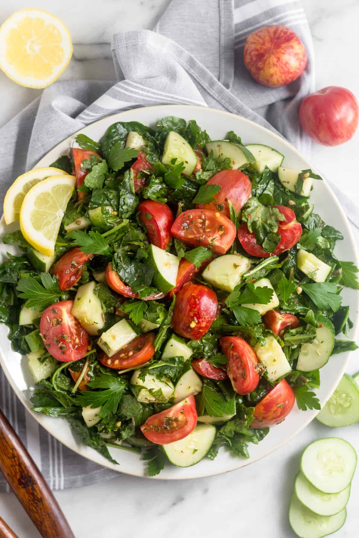 Large white plate filled with Chopped Tomato Cucumber Kale Salad surround by salad tongs, sliced cucumber, tomatoes, and half a lemon
