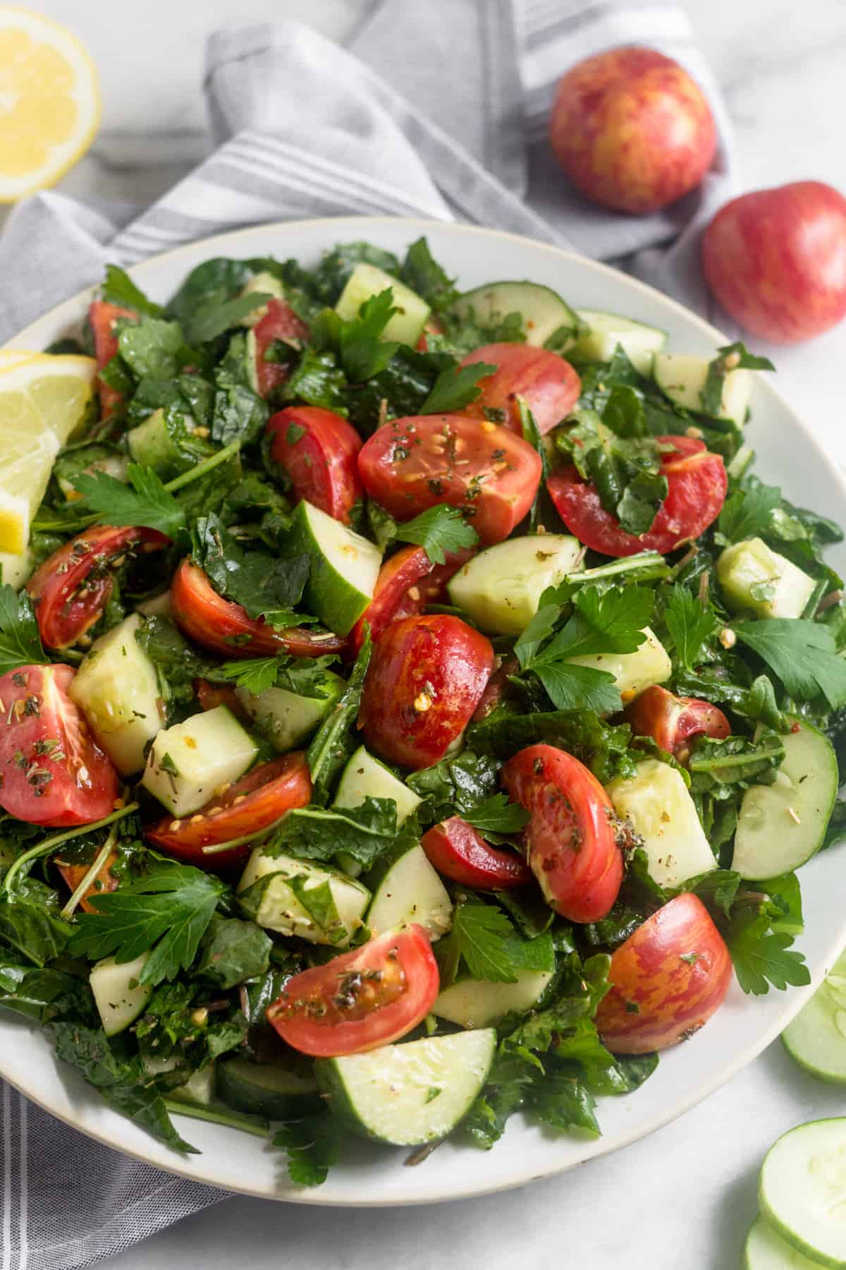 Chopped Tomato Cucumber Kale Salad on a large white plate with tomatoes, half a lemon, and sliced cucumber around it