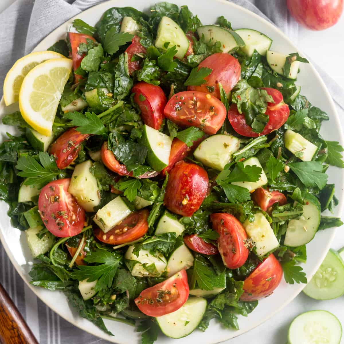 Large white plate filled with Chopped Tomato Cucumber Kale Salad surround by salad tongs, sliced cucumber, and tomatoes