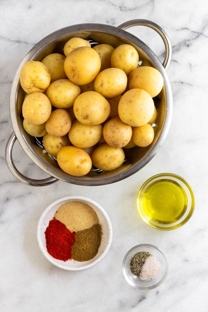 A marble counter with a colander of Yukon gold potatoes, a bowl of oil, a bowl of salt and pepper, and a bowl with 3 different spices in it.