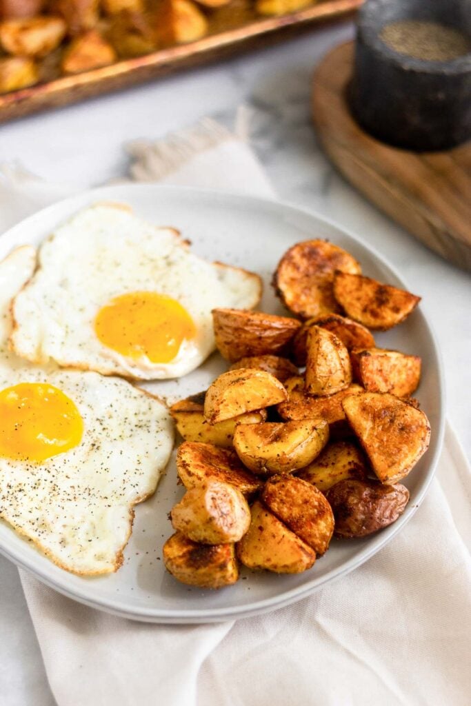 White plate with two fried eggs and roasted potatoes. Behind the plate is a pan of more potatoes and a small container filled with black pepper.