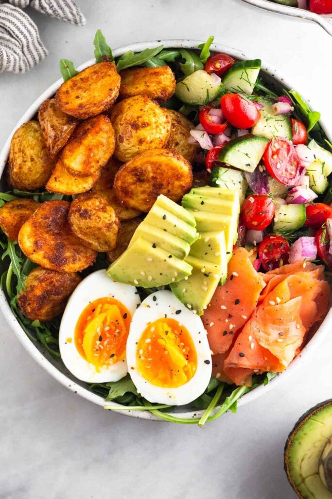 Breakfast bowl filled with greens, roasted potatoes, tomato and cucumber salad, smoked salmon, sliced avocado, and a hard-boiled egg and topped with everything but the bagel seasoning. Next to it is half an avocado.