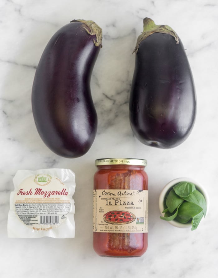 Countertop with 2 eggplants, mozzarella cheese, pizza sauce, and small bowl of basil.