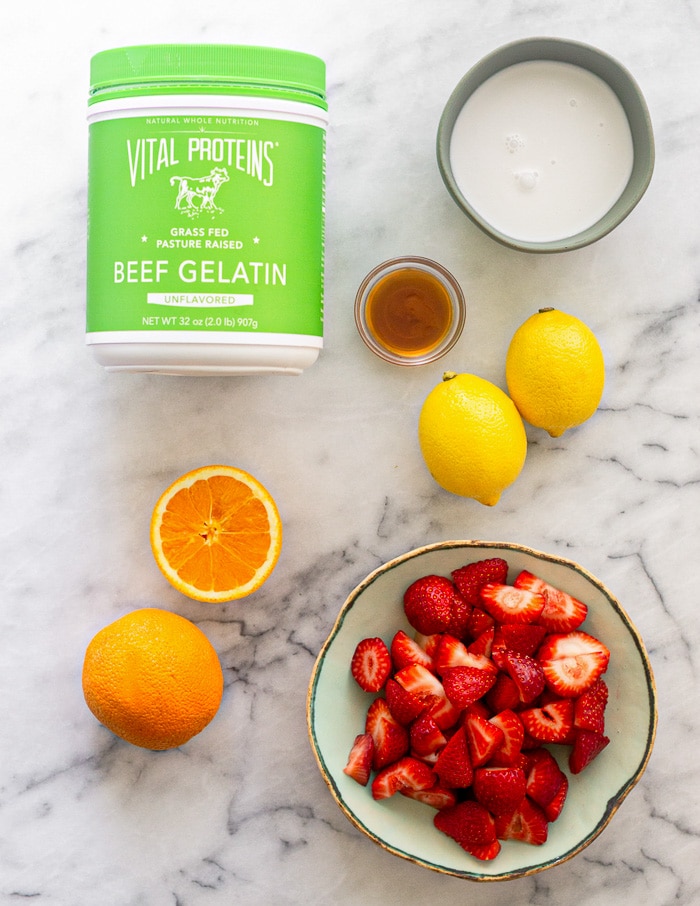 Overhead shot with a container of gelatin, bowl of coconut milk, lemons, bowl of honey, bowl of strawberries, and oranges.