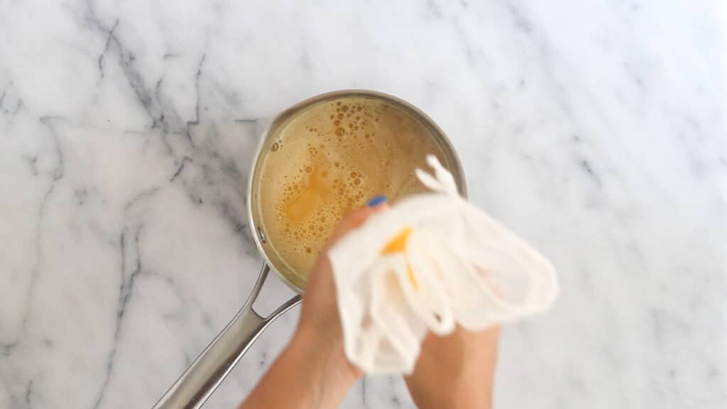 Overhead shot of a small sauce pan with fresh orange juice in it. Overtop a person is squeezing out fresh juice from a nut milk bag.