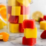 Gelatin gummies stacked on top of each other with a glass jar of them filled to the top behind them. There is also some fresh fruit spread out as well.