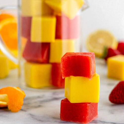 Gelatin gummies stacked on top of each other with a glass jar of them filled to the top behind them. There is also some fresh fruit spread out as well.
