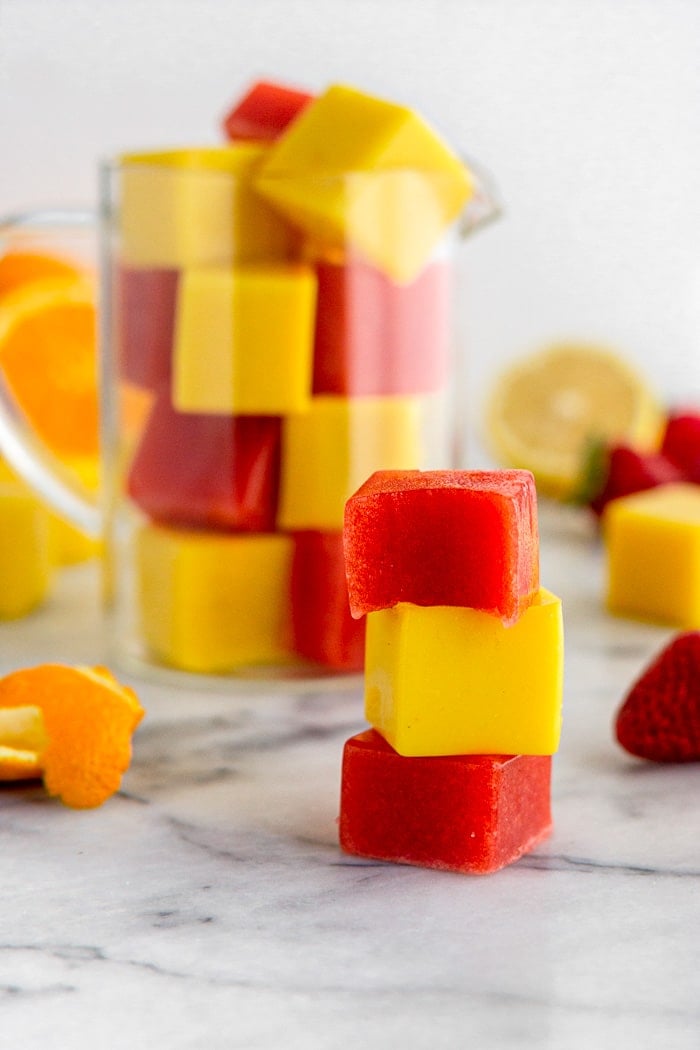 Gelatin gummies stacked on top of each other with a glass jar of them filled to the top behind them. There is also some fresh fruit spread out as well.