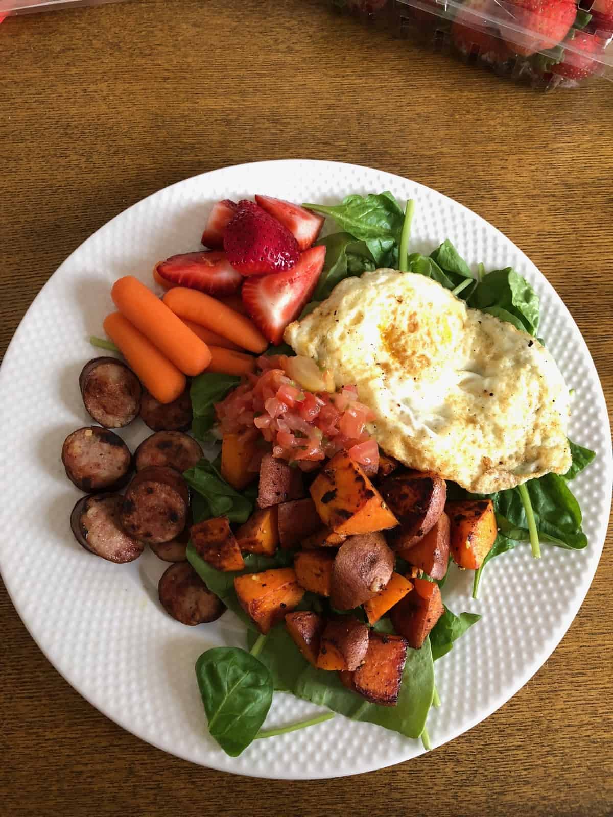 Breakfast plate of greens, fried egg, sweet potato, chicken sausage, carrots, strawberries, and salsa