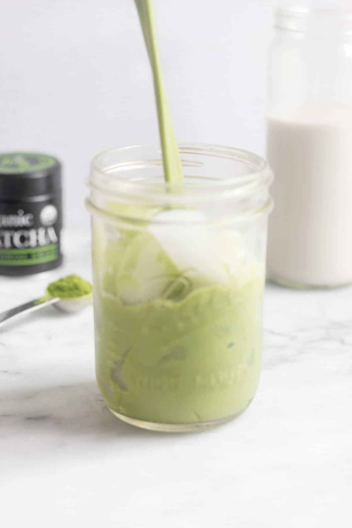 Glass jar with ice being filled with an iced matcha latte with a glass of almond milk and container of matcha behind it