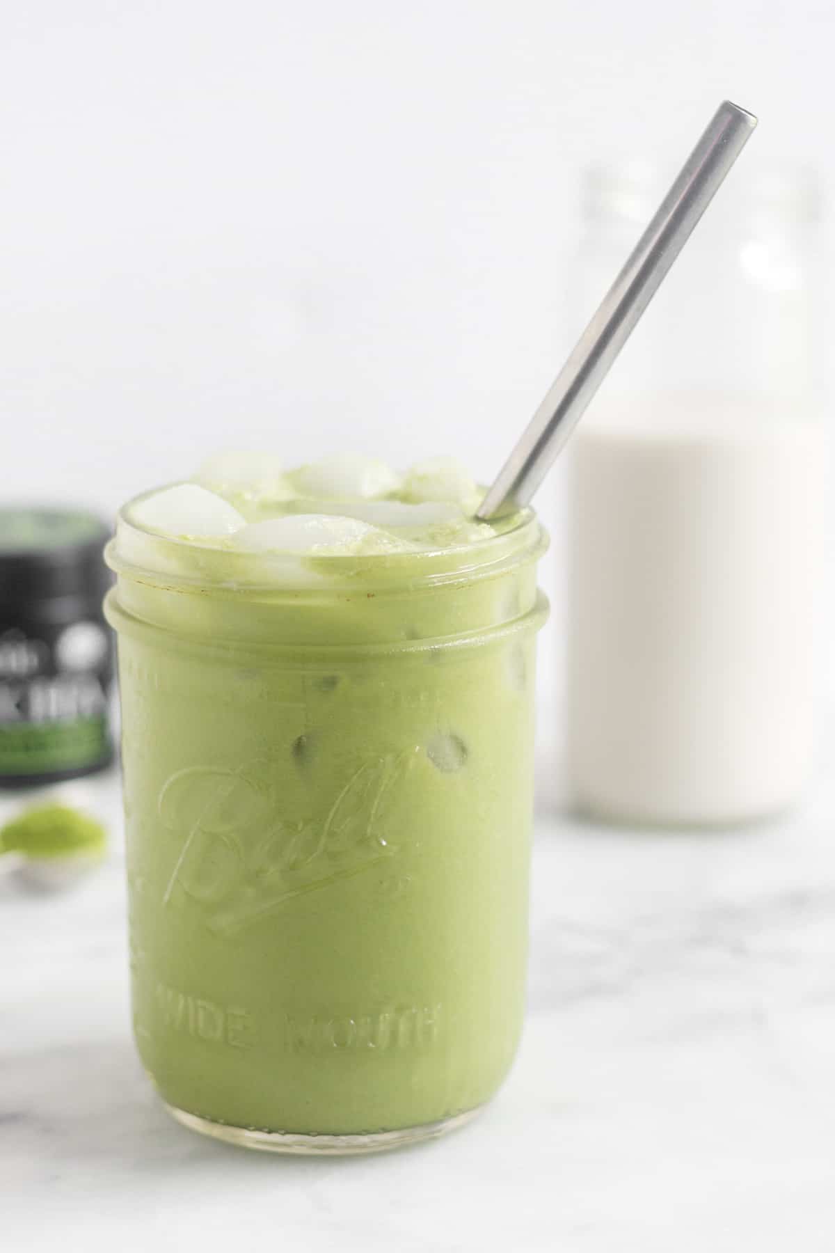 Glass jar with an iced matcha latte and a metal straw in it with a glass of almond milk and container of matcha behind it