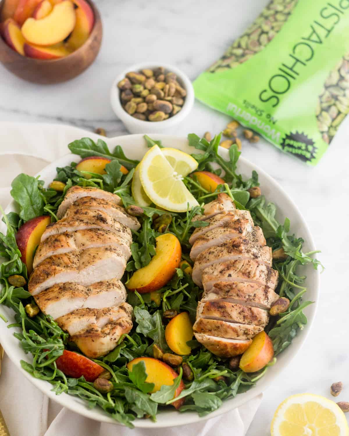 Peach and Pistachio Salad with Grilled Chicken salad in a large bowl with a bag of pistachios, a bowl of pistachios, and a bowl of cut up peaches behind it