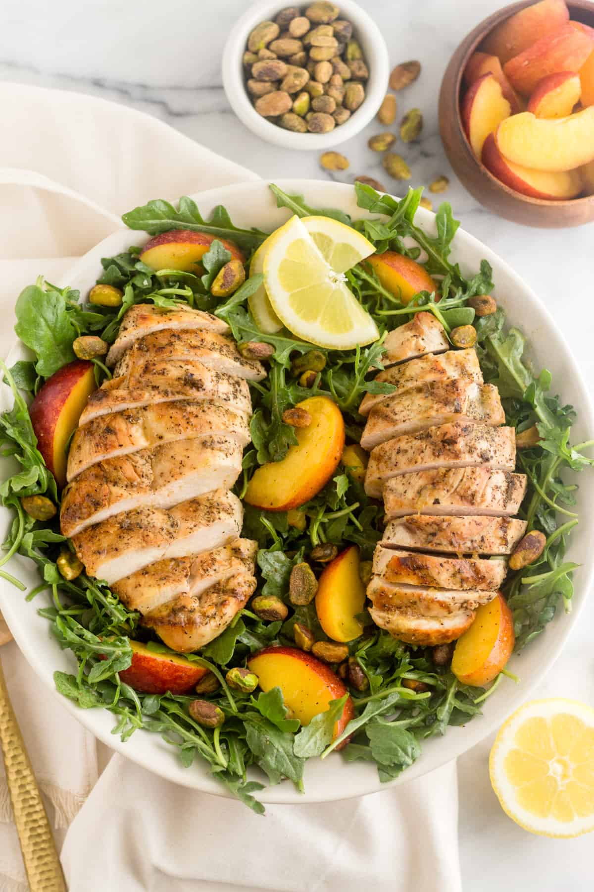 Peach and Pistachio Salad with Grilled Chicken in a large bowl overtop a white linen with pistachios, peaches, and half a lemon around it