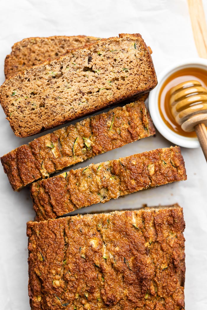 Overhead shot of sliced zucchini banana bread with the pieces spread out and some slices stacked on top of each other. Next to is a bowl of honey with a honey dipper.