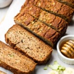 Healthy zucchini banana bread sliced on cutting board. Next to it is some shredded zucchini, sliced banana, and a bowl of honey.