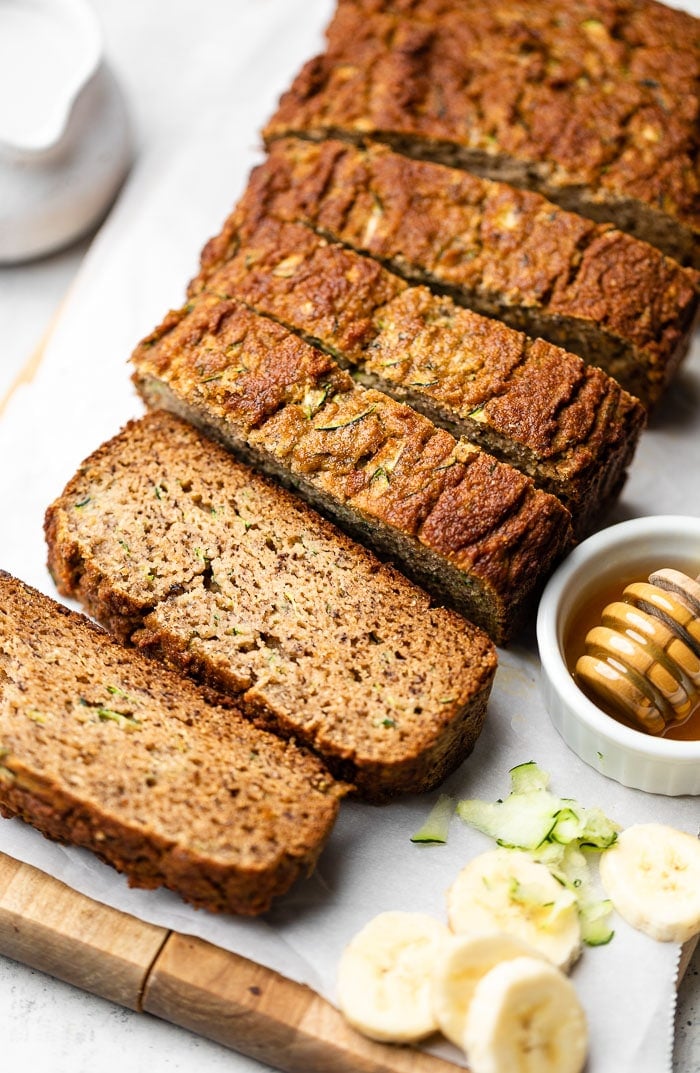 Healthy zucchini banana bread sliced on cutting board. Next to it is some shredded zucchini, sliced banana, and a bowl of honey.