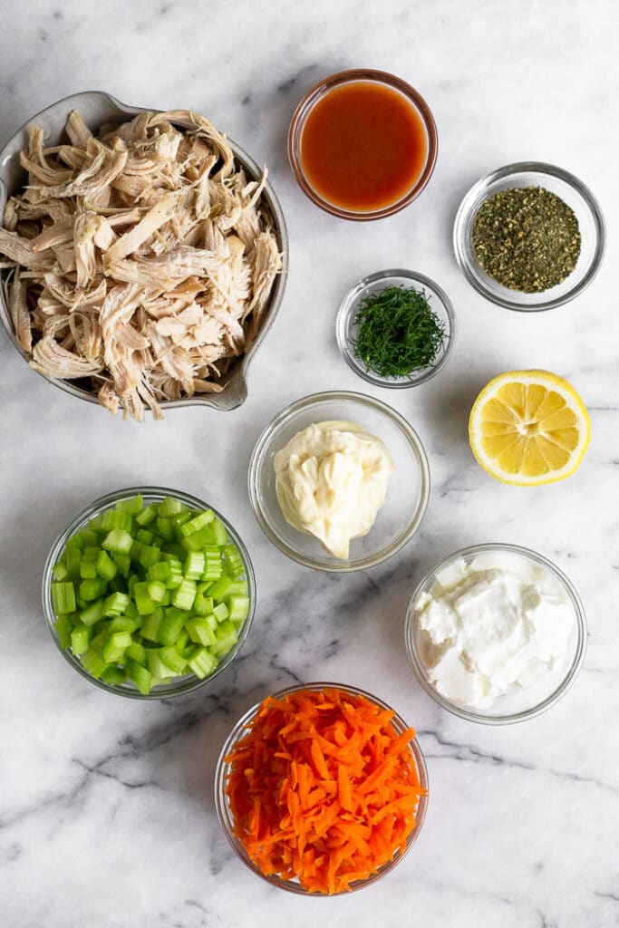 Overhead shot of a counter with a bowl of shredded chicken, bowl of hot sauce, bowl of ranch seasoning, bowl of dill, half a lemon, bowl of yogurt, bowl of shredded carrots, bowl of diced celery, and bowl of mayo.