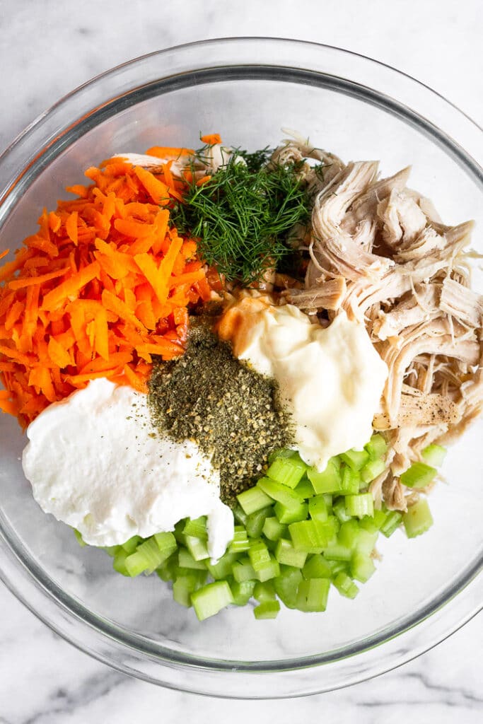 Large glass bowl with shredded chicken, mayo, diced celery, ranch seasoning, yogurt, shredded carrots, and dill.