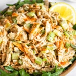 Buffalo chicken salad on top of some arugula in a bowl. It is topped with lemon wedges. In front of it is half a lemon and behind it is a plate of carrots and celery.
