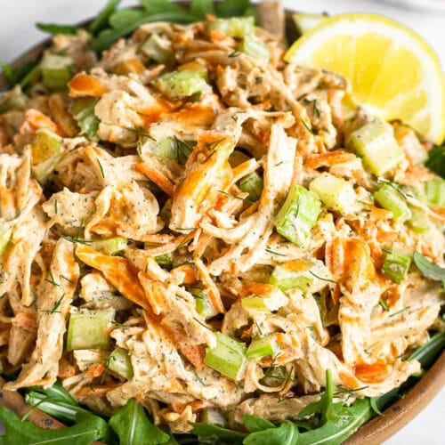 Buffalo chicken salad on top of some arugula in a bowl. It is topped with lemon wedges. In front of it is half a lemon and behind it is a plate of carrots and celery.