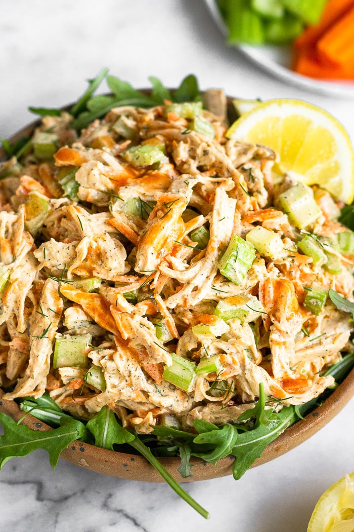 Buffalo chicken salad on top of some arugula in a bowl. It is topped with lemon wedges. In front of it is half a lemon and behind it is a plate of carrots and celery.